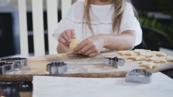 Klein meisje met lang haar in witte jurk maakt een snorren uit het ruwe deeg bij de voorbereiding van een koekjes van bakken. Kind maken van cookies van ruwe deeg in de vorm van sterren en harten — Stockvideo