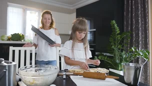 Menina pequena bonito fazendo biscoitos de massa crua na forma de corações e estrelas. Mulher bonita coloca bandeja de cozimento na mesa da cozinha. Filhinha com mãe assa biscoitos em casa no — Vídeo de Stock