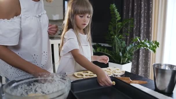 Young mother and little daughter making cookies in kitchen. Woman and little girl in kitchen placing biscuits dough on baking tray Stock Footage