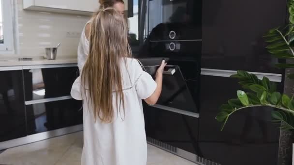 Visão traseira. Menina pequena com cabelos longos em vestido branco segurando uma bandeja de biscoito com biscoitos caseiros para assar. Linda mãe abre a porta do forno para biscoitos em casa. Mãe e filha fazendo biscoitos — Vídeo de Stock