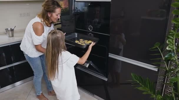 Niña en vestido blanco con pelo largo poner la bandeja para hornear con galletas en el horno. La joven y feliz familia está cocinando. Hornear — Vídeos de Stock