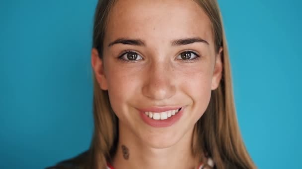 Close-up de um rosto bonito menina loira. A rapariga está a olhar para a câmara e a sorrir. Tiro de retrato. Menina adolescente atraente sorrindo para a câmera em ambientes fechados no fundo da parede azul — Vídeo de Stock