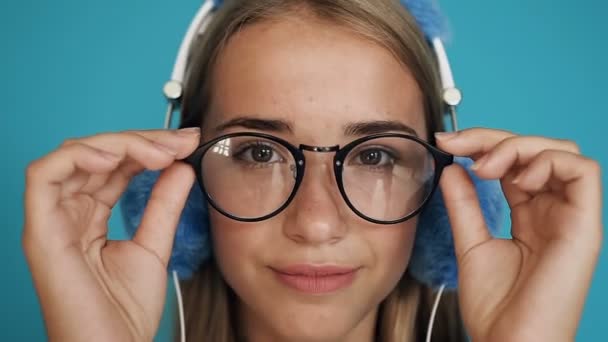 Portrait de la belle adolescente en lunettes et avec des écouteurs regardant droit dans l'appareil photo et souriant joyeusement sur le fond bleu ou turquoise. Ferme là. À l'intérieur — Video