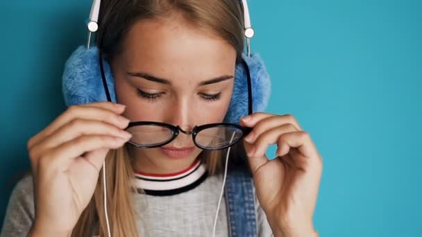 Retrato da menina bonita usando óculos e com fones de ouvido olhando diretamente na câmera e sorrindo alegremente no fundo da parede azul ou turquesa. Dentro de casa. Close-up — Vídeo de Stock