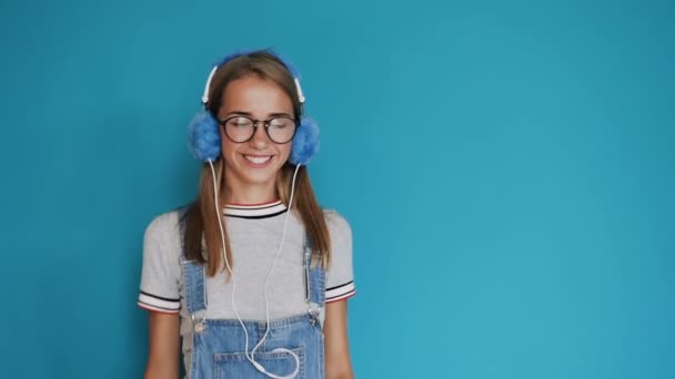 Attractive girl in glasses is dancing and moving to the rhythm listening music in headphones in indoors at blue background. Girl smiling and listening to music in headphones. Portrait of a beautiful — Stock Video