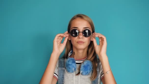 Adolescente modelo de moda menina vestindo óculos de sol modernos de forma redonda sorrindo olhando para a câmera, isolado no fundo da parede azul. Hipster menina usa óculos de sol e sorrindo. Estilo de beleza — Vídeo de Stock