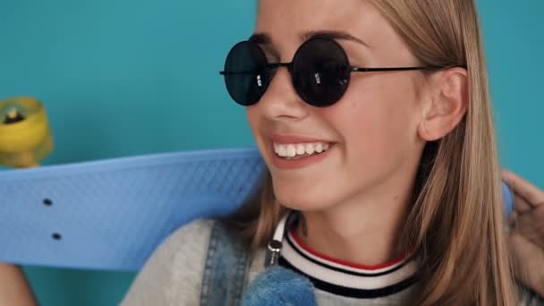 Портрет белой девушки в солнцезащитных очках с улыбкой на камере со скейтбордом в руках на синем фоне. Крупный план хипстера молодая девушка улыбается со скейтбордом — стоковое видео