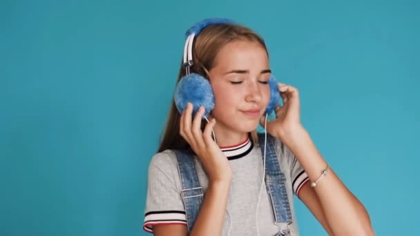 Krásná usměvavá dívka má na sobě velké modré sluchátka poslouchat hudbu uvnitř studio. Ruce na sluchátka. Roztomilý dospívající dívka poslechu hudby přes sluchátka na modrém pozadí — Stock video