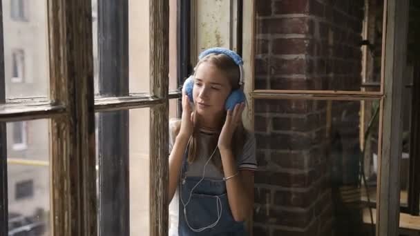 Retrato de niña atractiva en el fondo de la ventana escuchando música con auriculares. Una hermosa chica se sienta cerca de una ventana con auriculares escuchando música desde el teléfono inteligente — Vídeos de Stock