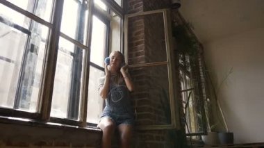 Rahatlatıcı beyaz kız tuğla duvara yakın pencere kenarında oturan ve onun kulaklık müzik. Çekici genç hippi kız mavi kulaklık aracılığıyla müzik dinlemek