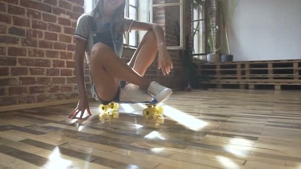 Lebensstil. Porträt eines attraktiven blonden Teenie-Mädchens in Jeans-Overalls mit langen Haaren sitzt auf einem Skateboard und blickt lächelnd in die Kamera. Niedliche Skateboarder Mädchen sitzt auf dem Skateboard in — Stockvideo