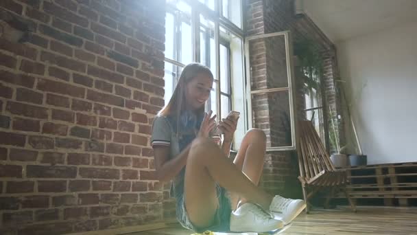 Девушка-хипстер, сидящая на скейтборде, улыбается и использует смартфон в здании на кирпичной стене и фоне окна. Девушка-хипстер со смартфоном, наушники на скейтборде — стоковое видео