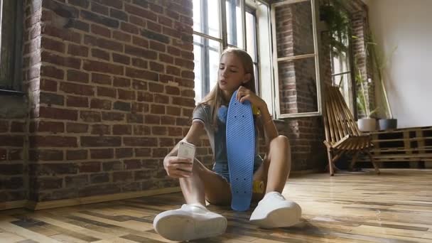 Adolescente attrayante est assise sur le plancher tenant skateboard et fait des photos selfie sur son smartphone à l'intérieur. Belle fille hipster avec planche à roulettes prenant selfies au mur de briques rouges et — Video