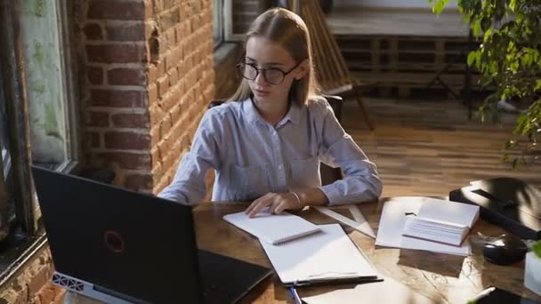 Стильна біла молода бізнес-леді, використовуючи ноутбук, сидить за столом в модному офісному червоному цегляному інтер'єрі. Дівчина-підліток з довгим волоссям в окулярах навчається через ноутбук і пише записку на ноутбук — стокове відео
