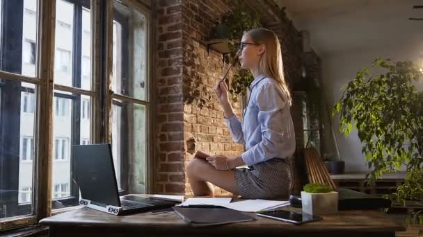 Hermosa chica caucásica atractiva se sienta en la mesa con un bloc de notas y pluma en las manos. Personalidad creativa se sienta sobre la mesa con un cuaderno en el interior moderno — Vídeos de Stock