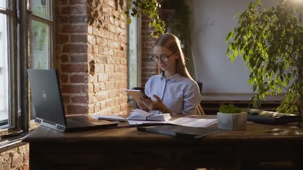 Succesvolle jonge zakenvrouw in glazen met lange haren maakt berekeningen op haar werkplek met behulp van tablet pc. Met behulp van een laptop, Tablet PC- en documenten werken bij moderne zakenvrouw — Stockvideo