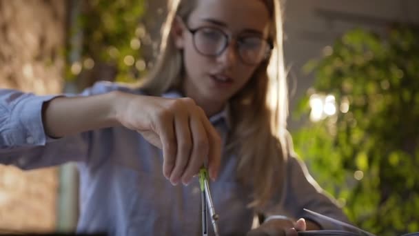 Close-up de uma bela menina estudante em óculos sentados ao lado da mesa usa a bússola em uma folha de papel, ela desenha um círculo. Mulher de negócios fazendo um relatório sobre o marketing de sucesso. Visão de — Vídeo de Stock