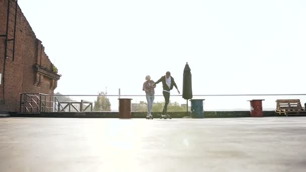 Красивая молодая пара держится за руки, катаясь на скейтборде на крыше современного здания. Мальчик и девочка катаются на скейтборде на крыше на открытом воздухе Концепция современного городского активного образа жизни — стоковое видео