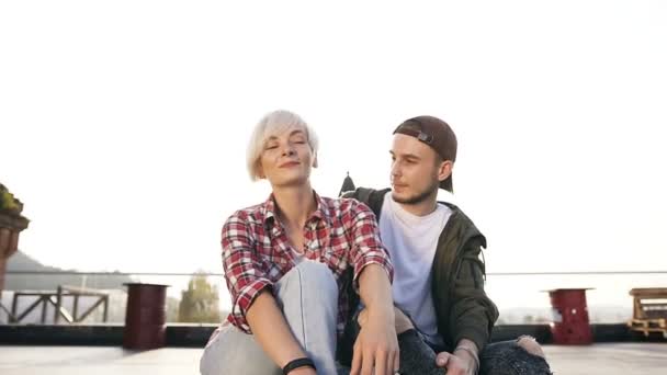Két fiatal szép pár ül együtt egy hosszú gördeszka a tetőn, egy modern épület. Egy fiatal srác és a lány lovagolni a gördeszka a naplemente egy nyári napon — Stock videók