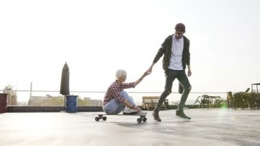 Yakışıklı hipster adam sevgilisini kaykaylar modern binanın çatı, eğleniyor. Şık çift longboard modern binanın çatısına kaykay ile eğleniyor