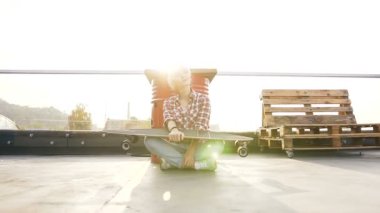 Kot pantolon ve kaykay holding endüstriyel bina modern çatı katta oturan kırmızı kareli gömlek giyen genç hippi kız. Açık havada uzun ile yaz gününde oturan sarışın kız