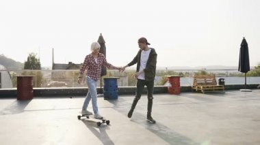 Yakışıklı olan mekanlarda kaykay üzerinde sürme için kız arkadaşı öğretir. Hipster genç adam ve kız var eğlenceli gün batımında modern bina çatı üzerinde kaykay sürme