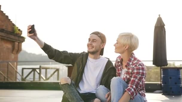 Güzel şık çift longboard kaykay üzerinde çatı bahçesi, endüstriyel bina ve selfie fotoğraf üstünde smartphone yapım üzerinde oturuyorsun. Genç adam ve kız eğleniyor yapıyorsun — Stok video