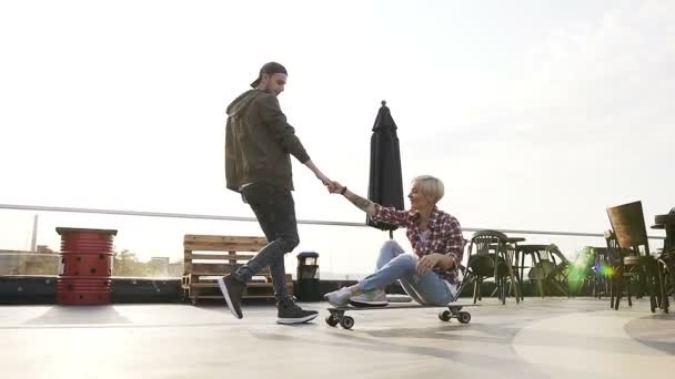 Hermosa pareja joven se divierten en el skateboard al aire libre en la azotea del edificio. Caucásico joven mujer está sentado en un longboard con el apoyo de su novio — Vídeo de stock