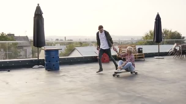 英俊的小伙子和他的女孩在工业大厦屋顶的滑板上玩得很开心。两个人玩长板滑板在现代建筑的屋顶。现代概念 — 图库视频影像