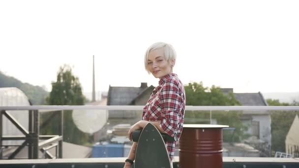 Retrato de menina hipster loira atraente com um skate posando para a câmera e sorrindo ao ar livre. Bonito jovem mulher em xadrez camisa vermelha está de pé no telhado do edifício moderno com — Vídeo de Stock