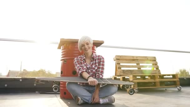 Pretty blonde girl sits on the floor with longboard outdoors at summer sunset. Young Caucasian skater wearing jeans and red checkered shirt holding long skate board on modern rooftop of building — Stock Video