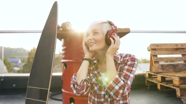 Linda chica rubia en auriculares rojos escuchando música y bailando. Joven mujer hipster con auriculares bailando, sonriendo y agitando su cabeza al aire libre al atardecer — Vídeo de stock