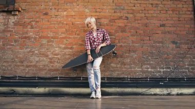 Gün batımında kırmızı tuğla duvar yakınındaki çatıda longboard konumu olan genç sarışın kız. Güzel sarışın kız longboard, tuğla duvar, çatı arka plan ile modern bina
