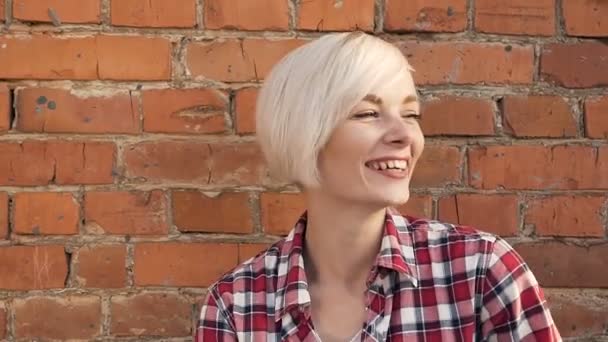 Elegante modelo chica rubia está de pie junto a la pared de ladrillo rojo. Retrato de una joven hermosa mujer rubia caucásica sonriendo al aire libre. Primer plano de una joven feliz — Vídeos de Stock