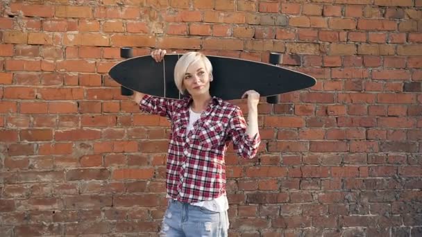 Porträt einer kaukasischen jungen Frau, die mit einem langen Skateboard in den Händen vor rotem Backsteinhintergrund posiert und in die Kamera lächelt. schönes blondes Mädchen mit Kurzhaarschnitt hält lang — Stockvideo