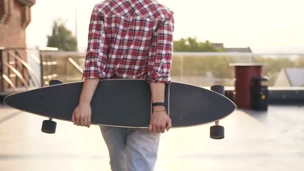 A menina bonita vai em uma rua com um skate em mãos. Uma visão traseira de uma jovem mulher andando segurando um skate em suas mãos no telhado do edifício moderno contra o fundo da cidade — Vídeo de Stock