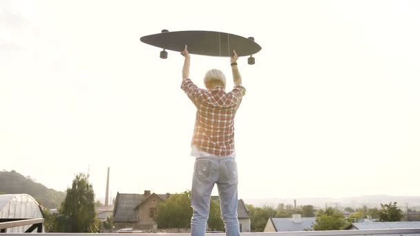 Vista trasera, joven mujer hipster con monopatín está de pie en la azotea moderno skatepark en la hora de verano al atardecer. Niña sosteniendo el monopatín largo está de pie en lo alto del techo del edificio moderno y — Vídeos de Stock