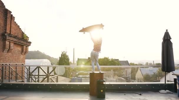 Visão traseira linda menina loira em camisa xadrez e jeans com skate longo em suas mãos no telhado ao pôr do sol. Uma menina com corte de cabelo curto está dançando com um skate em suas mãos no — Vídeo de Stock