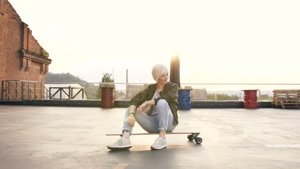 Cute teen girl is sitting on her skateboard of the rooftop modern building. Young blonde woman sitting on longboarding at sunrise at the city — Stock Video