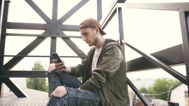 Joven hombre hipster guapo usando teléfono inteligente en las escaleras de hierro negro del edificio industrial al atardecer. joven usando el teléfono inteligente mientras está sentado en las escaleras fuera de la ciudad — Vídeos de Stock