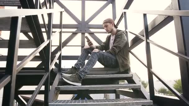 Jeune homme beau avec smartphone assis à des escaliers en fer. Teenage hipster boy en utilisant smartphone wifi connecté, concept moderne de banlieue de style de vie — Video