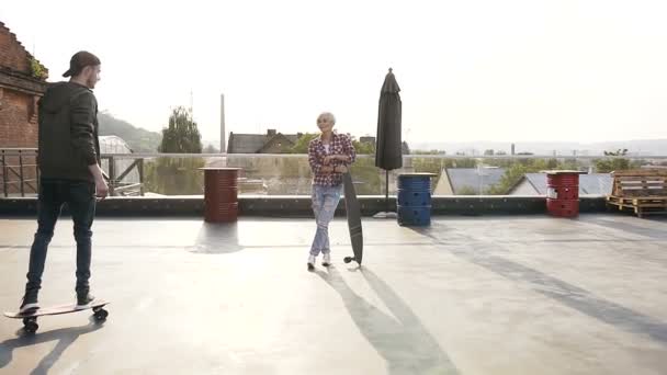 Bonito casal jovem estão se divertindo com skates no telhado do edifício moderno ao pôr do sol. Dois amigos andam de skate. Conceito de estilo de vida urbano moderno — Vídeo de Stock