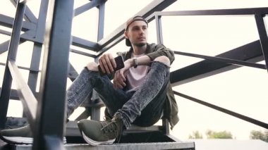 Elinde bir endüstriyel günbatımında bina merdivenlerde oturan smartphone yakışıklı adam. Siyah demir merdivenlerde genç hipster adam oturur