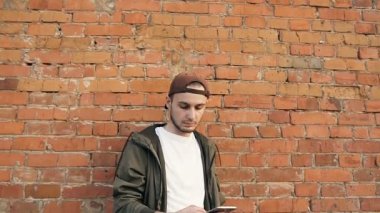 Kentsel şehir arka planda tuğla duvar üzerinde rahatlatıcı Smartphone kullanan genç yakışıklı adam. Mutlu hipster adam mobil oyunlar oynarken. Açık havada