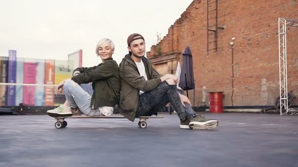 Os amigos estão se divertindo enquanto estão sentados em um skate ao ar livre. Jovem casal hipster se divertindo com skate longo no telhado de um edifício moderno — Vídeo de Stock