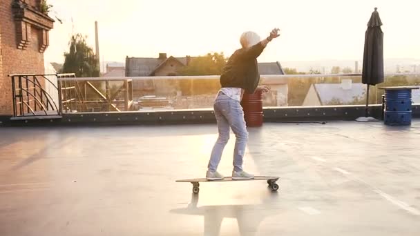 Hermosa chica monta en un longboard. Chica Hipster en camisa de chaqueta y vaqueros azules paseos en un longboard en el techo de un edificio moderno. Al aire libre — Vídeo de stock