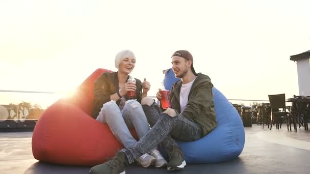 Joven pareja caucásica enamorada sentada en sillones multicolores en la terraza de la cafetería, bebiendo jugos. Guapo g uy y novia hablando entre sí en la terraza en la cafetería al atardecer. Al aire libre — Vídeo de stock