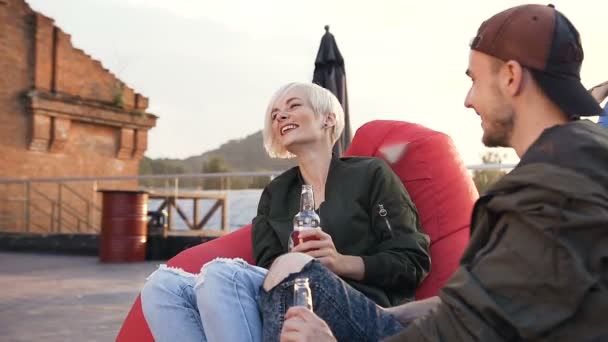 Joven y chica están sentados en sillones multicolores en la terraza café beber bebida energética. Dos amigos divirtiéndose beber cerveza — Vídeos de Stock