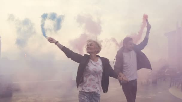 Casal feliz correndo com fumaça colorida no terraço do edifício moderno. Jovem casal hipster com bombas de fumaça coloridas ao pôr do sol. Ao ar livre — Vídeo de Stock