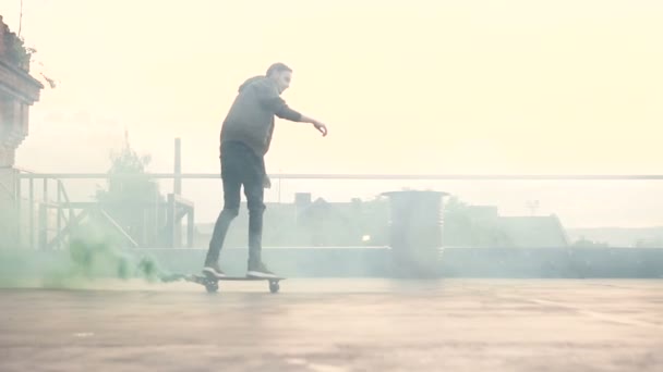 Bonito homem jovem passeios de skate com fumaça verde no terraço do edifício industrial. Skateboarder fazendo uma acrobacia em seu skate no telhado do edifício industrial — Vídeo de Stock